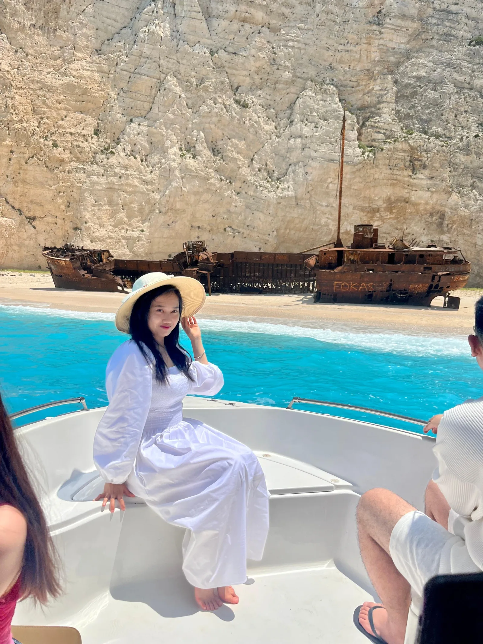 A woman in a white dress on the bow of the boat with the rusted Panagiotis wreck and the beach behind