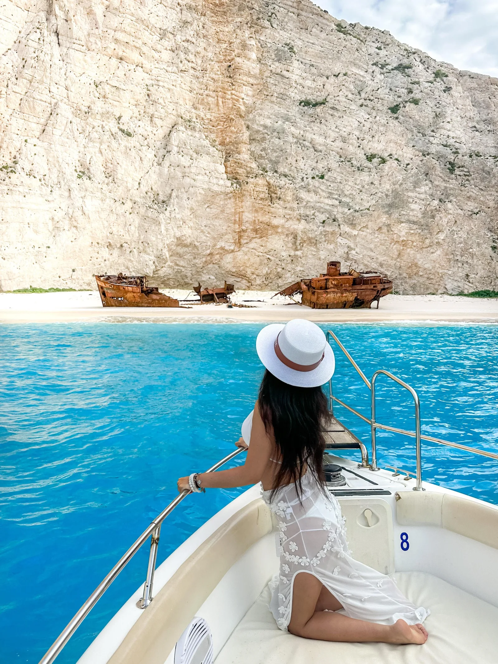 A passenger in a white hat on the bow of the speedboat with the wreck and beach behind