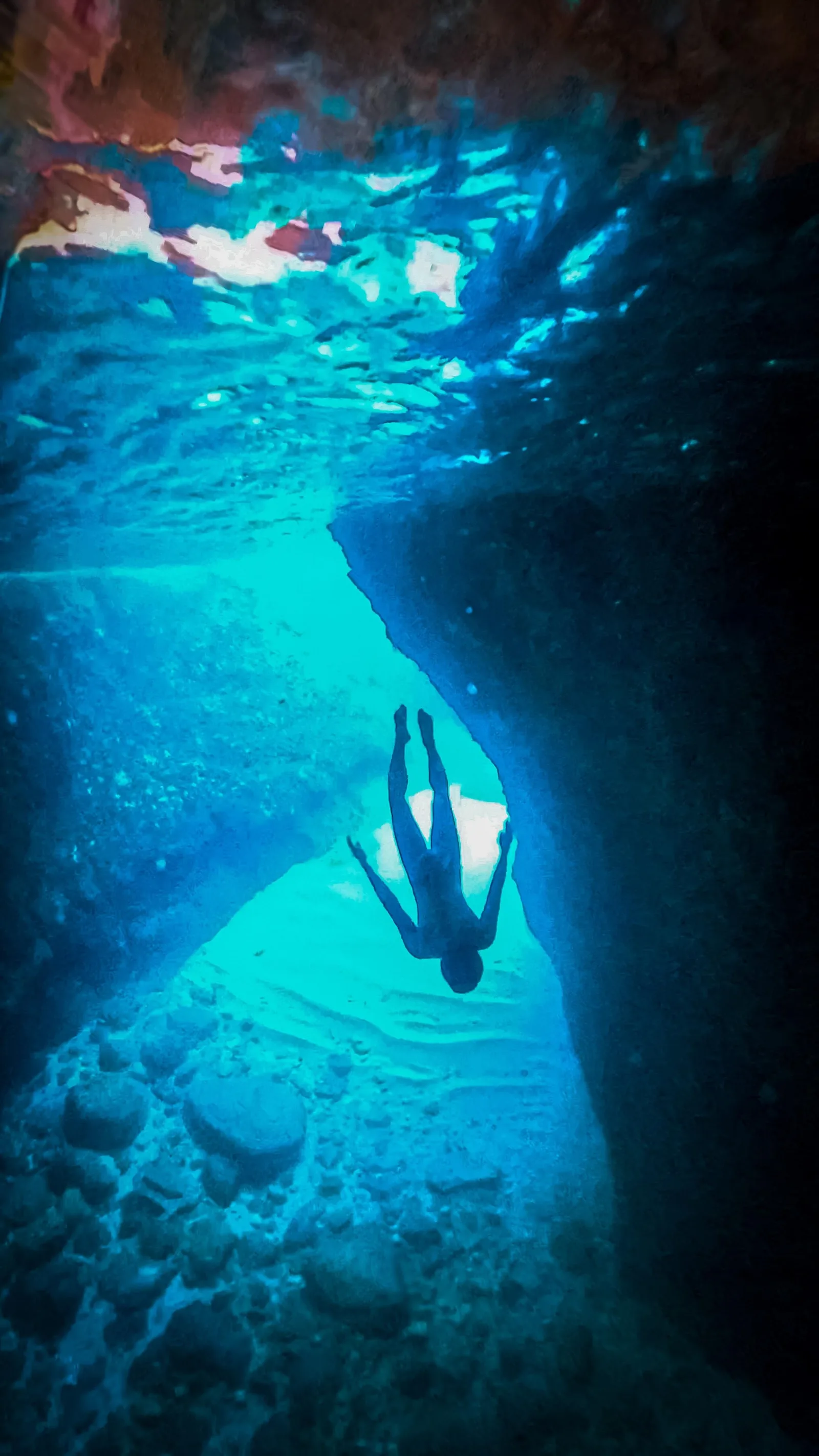 A snorkeller swimming through a cave arch lit turquoise from below